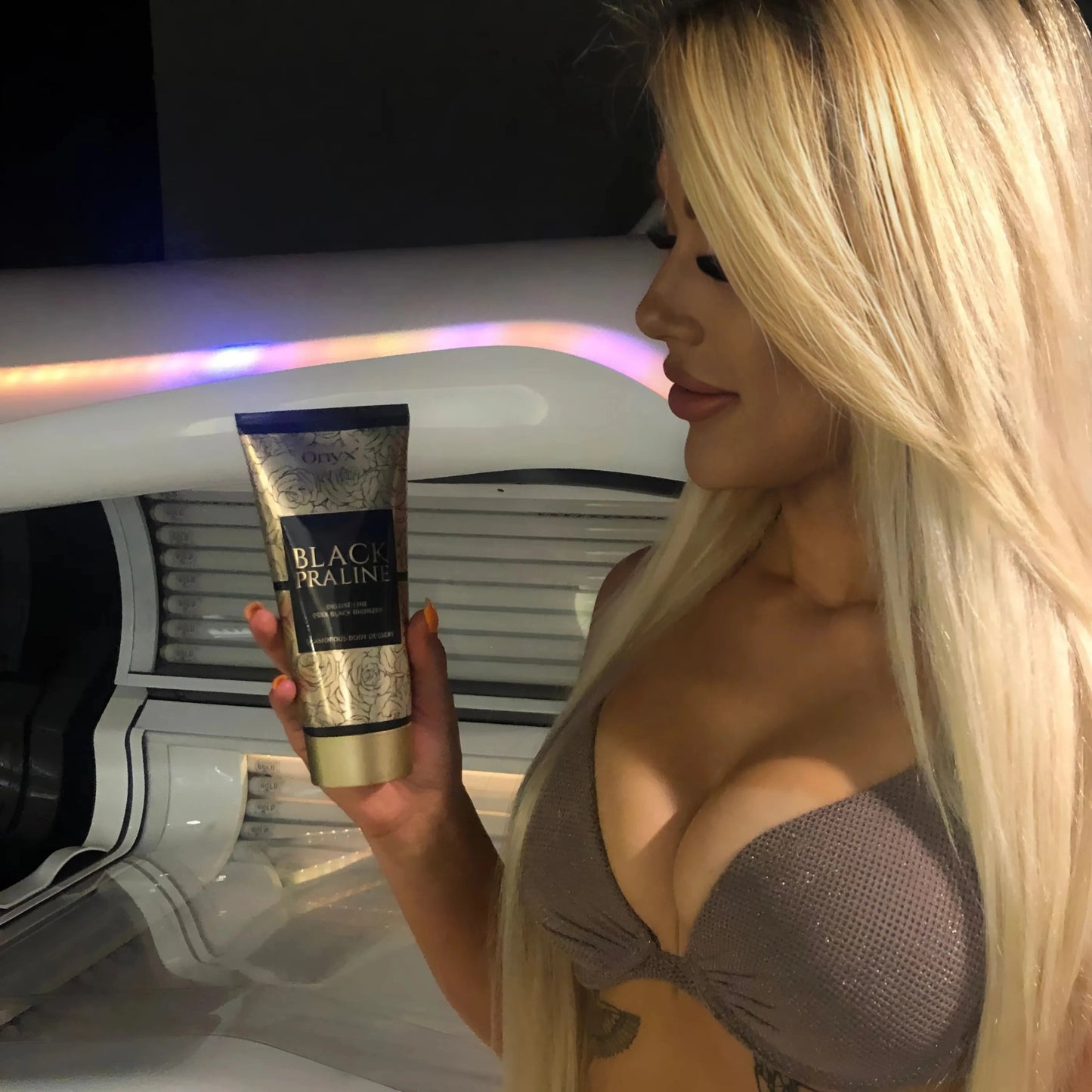 Mujer rubia de cabello largo sostiene la loción Onyx Black Praline frente a un solarium y la observa