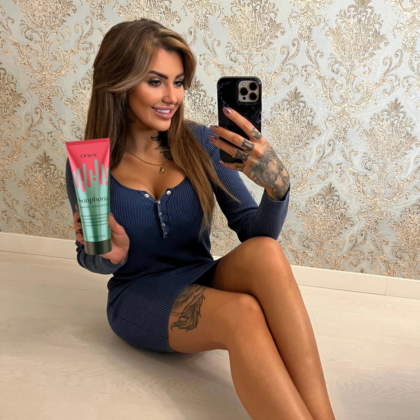 Mujer con tatuajes sostiene la Loción Bronceadora Onyx Sunphoria en un selfie frente al espejo