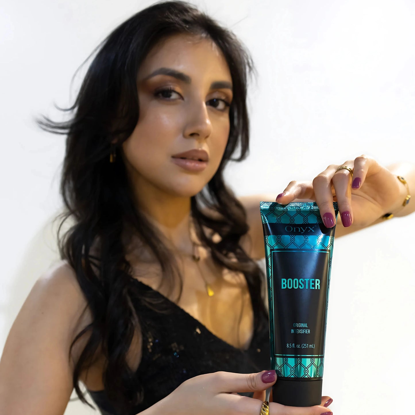 Mujer con vestido negro presenta la Loción Bronceadora Onyx Tanning Booster con ambas manos sobre fondo blanco – elegante momento de belleza