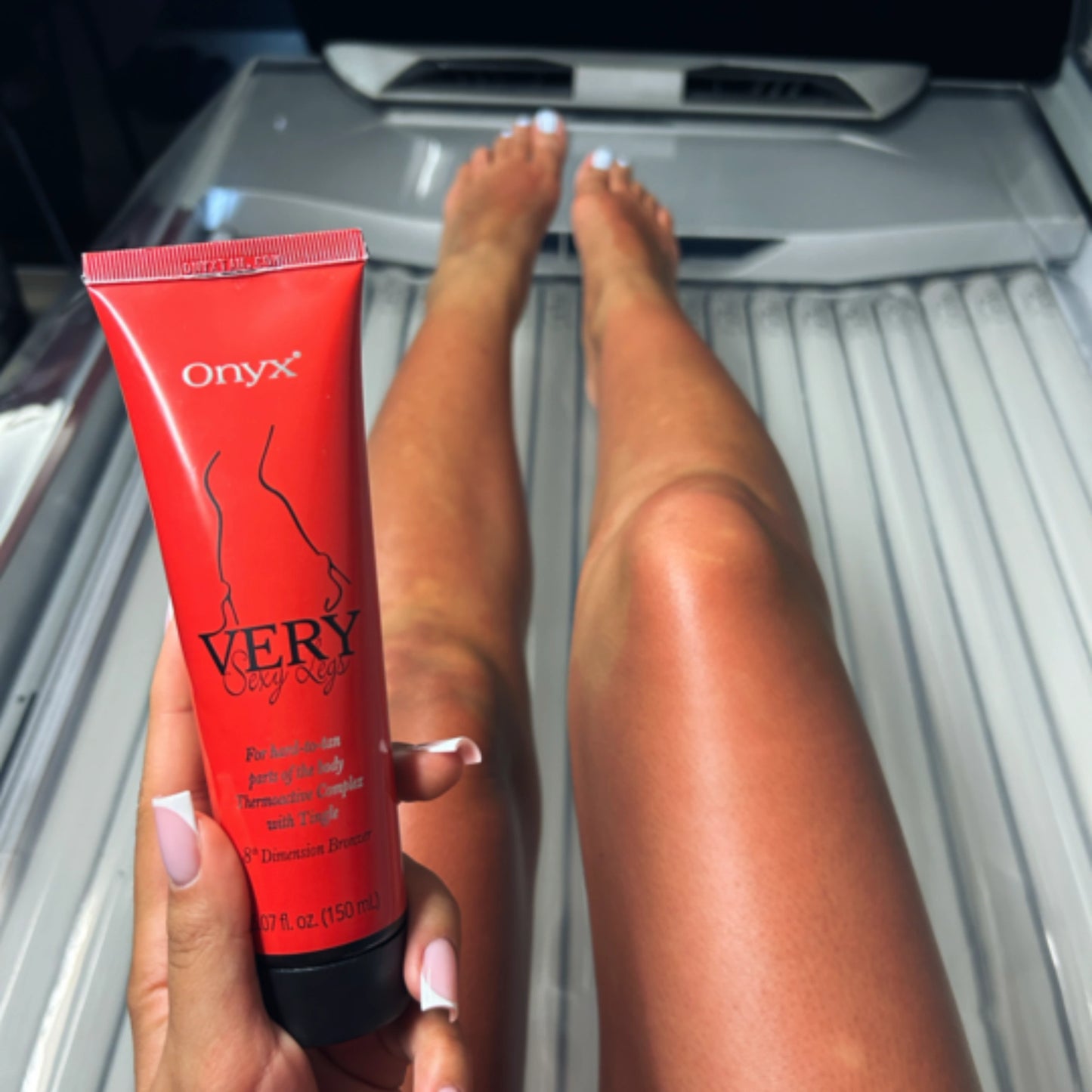 Piernas bronceadas de una mujer tras aplicar Onyx Very Sexy Legs en el solárium, sosteniendo la loción en la mano