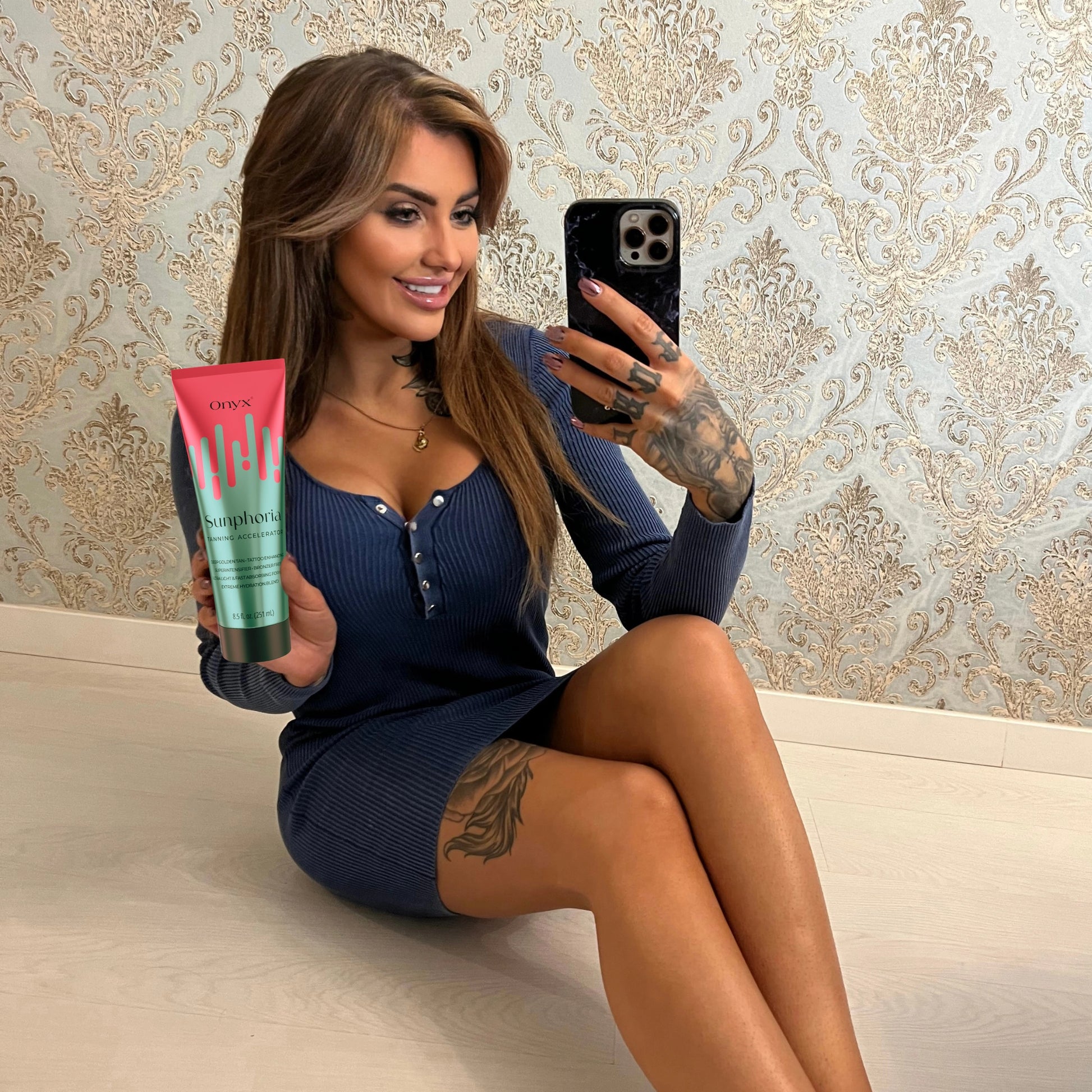 Mujer con tatuajes sostiene la Loción Bronceadora Onyx Sunphoria en un selfie frente al espejo