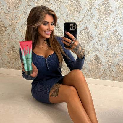 Mujer con tatuajes sostiene la Loción Bronceadora Onyx Sunphoria en un selfie frente al espejo