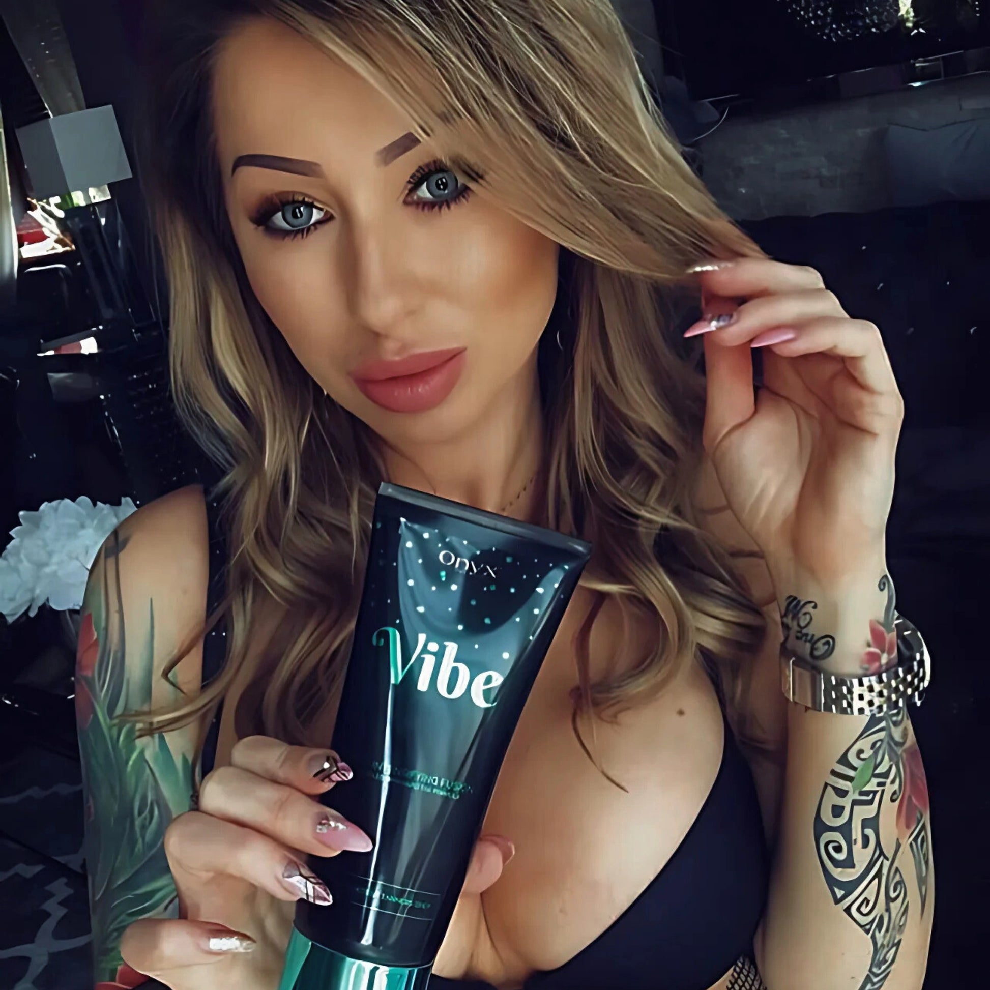 Mujer con tatuajes sostiene la Loción Bronceadora Onyx Vibe en la mano – cuidado específico para piel bronceada con estilo individual